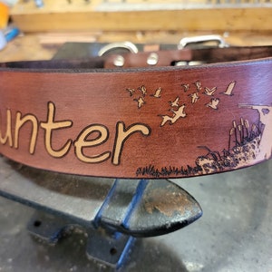 May include: Brown leather dog collar with the word "Hunter" engraved on it. The collar also features an engraved design of a hunter with a shotgun and a flock of birds flying overhead.