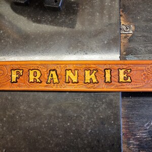 May include: A brown leather belt with a floral design and the name "FRANKIE" embossed in gold lettering.