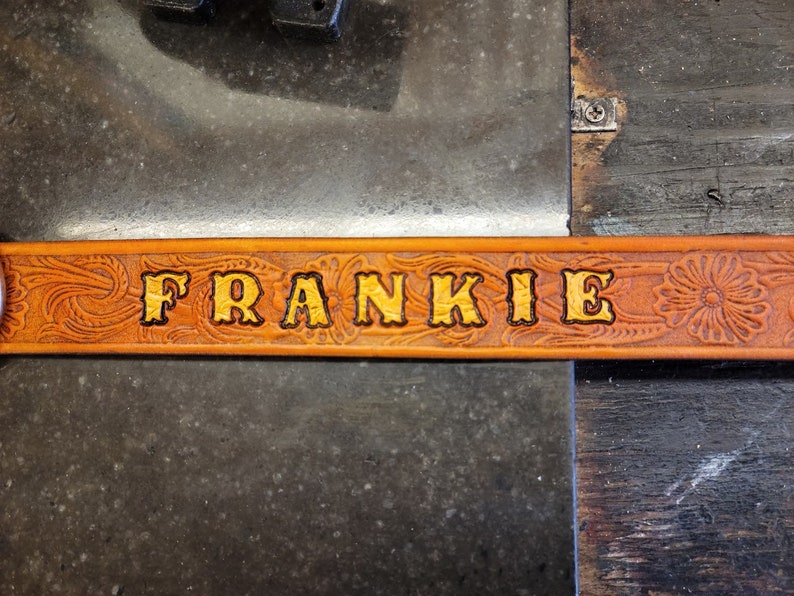 May include: A brown leather belt with a floral design and the name "FRANKIE" embossed in gold lettering.