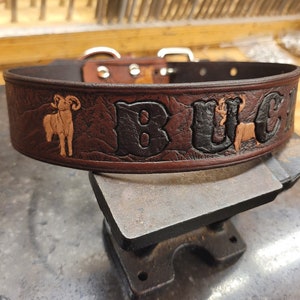 May include: A brown leather dog collar with the name "BUCK" embossed in black lettering. The collar also features two brown, embossed images of a ram and a deer.