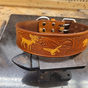 May include: A brown leather dog collar with a yellow embossed design of a deer, a duck, and a bear in a forest setting.