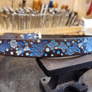 Puede incluir: Un cintur&oacute;n de cuero marr&oacute;n con un dise&ntilde;o floral azul y blanco. El cintur&oacute;n est&aacute; decorado con peque&ntilde;os puntos blancos y flores azules con centros blancos. El cintur&oacute;n est&aacute; sobre una superficie de trabajo de cuero marr&oacute;n.