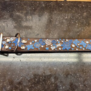 Puede incluir: Un collar de perro de cuero marr&oacute;n con un dise&ntilde;o floral azul y blanco. El collar tiene detalles met&aacute;licos plateados y una hebilla.