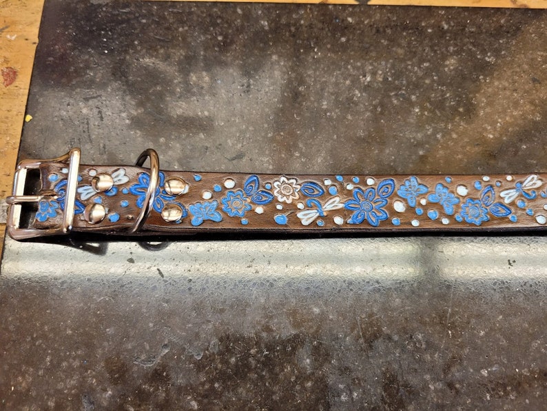 Puede incluir: Un collar de perro de cuero marr&oacute;n con un dise&ntilde;o floral azul y blanco. El collar tiene detalles met&aacute;licos plateados y una hebilla.