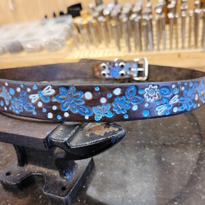 Puede incluir: Un cintur&oacute;n de cuero marr&oacute;n con un dise&ntilde;o floral azul y blanco. El cintur&oacute;n tiene una hebilla plateada y est&aacute; sobre una superficie de metal negro.