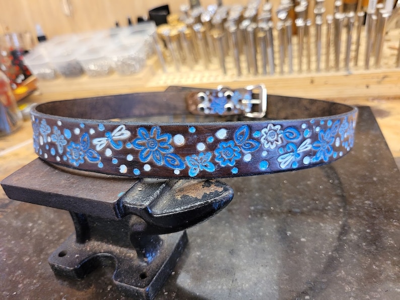Puede incluir: Un cintur&oacute;n de cuero marr&oacute;n con un dise&ntilde;o floral azul y blanco. El cintur&oacute;n tiene una hebilla plateada y est&aacute; sobre una superficie de metal negro.