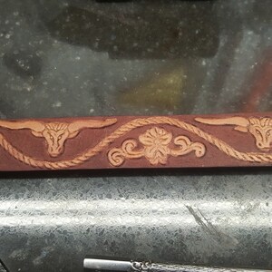 May include: A brown leather belt with a decorative design featuring a repeating pattern of a rope, a stylized flower, and a bull's head.