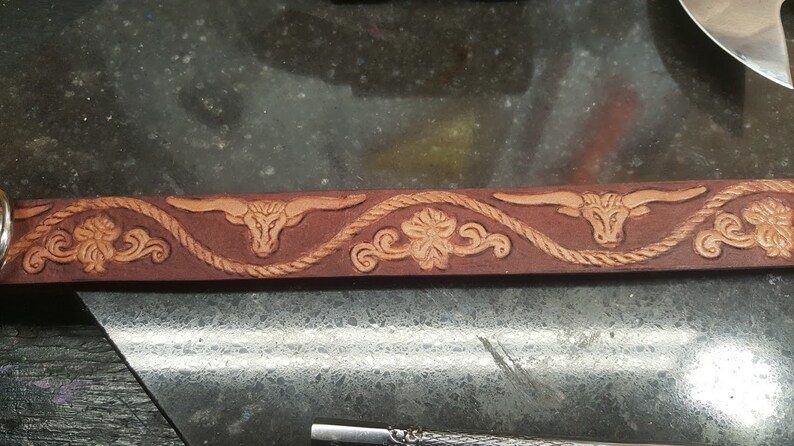 May include: A brown leather belt with a decorative design featuring a repeating pattern of a rope, a stylized flower, and a bull's head.