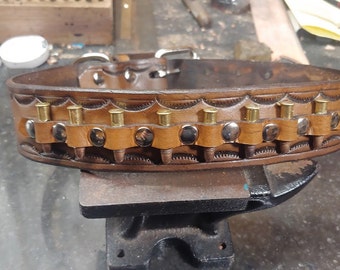 May include: A brown leather belt with a decorative design and brass bullet casings. The belt has silver studs and is on a black metal surface.