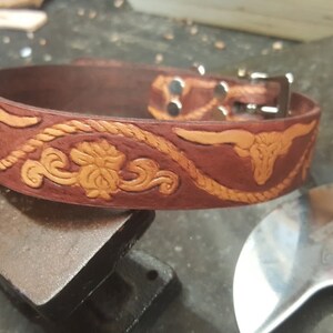 May include: A brown leather dog collar with a tooled design of a rope, floral patterns, and a bull's head. The collar has a silver buckle and two silver rivets.