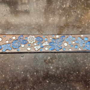 Puede incluir: Un cintur&oacute;n de cuero marr&oacute;n con un dise&ntilde;o floral blanco y azul. El dise&ntilde;o incluye mariposas, flores y lib&eacute;lulas.