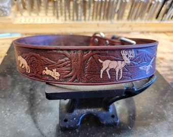 Puede incluir: Un collar de perro de cuero marrón con un diseño en relieve detallado de un ciervo, un conejo y un pájaro en un entorno de bosque.