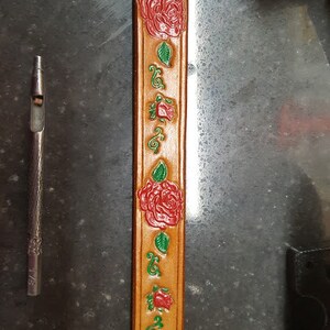 May include: A close-up of a brown leather strap with a repeating pattern of red roses and green leaves. The strap is decorated with a floral design.