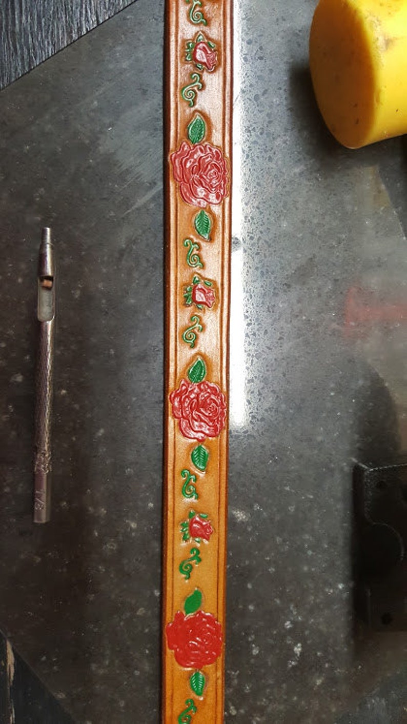 May include: A close-up of a brown leather strap with a repeating pattern of red roses and green leaves. The strap is decorated with a floral design.
