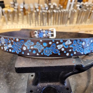 Puede incluir: Un cintur&oacute;n de cuero marr&oacute;n con un dise&ntilde;o floral azul y blanco. El cintur&oacute;n est&aacute; sobre un yunque de metal.