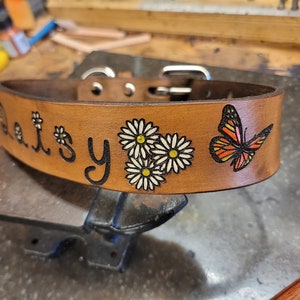 May include: A brown leather dog collar with the name "Daisy" engraved on it. The collar is decorated with three white daisies and a colorful butterfly.