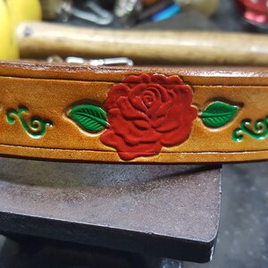 May include: A brown leather belt with a red rose and green leaves design. The belt is decorated with a floral pattern.