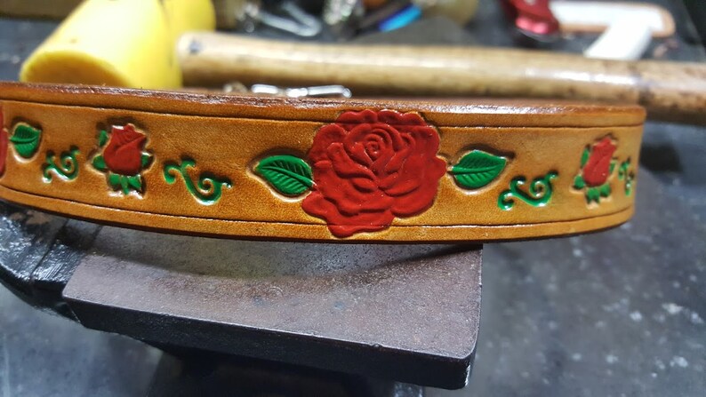May include: A brown leather belt with a red rose and green leaves design. The belt is decorated with a floral pattern.