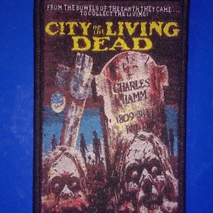 May include: A black and red embroidered patch featuring the text "City of the Living Dead" and a graphic of a zombie head rising from the ground. The text "From the bowels of the earth they came... to collect the living!" is also included.