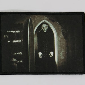 Puede incluir: Un parche bordado en blanco y negro que representa a un hombre con un traje oscuro de pie en una puerta. El hombre tiene la piel pálida y una cara demacrada. El fondo es oscuro y sombrío.
