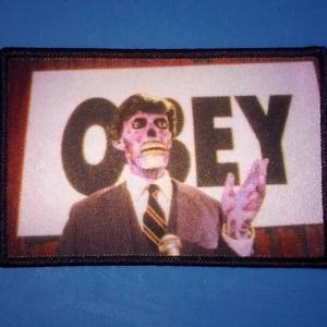 May include: A black and white patch with a man in a suit and a microphone. The man has a pale face and a large head. The text "OBEY" is in large black letters behind him.