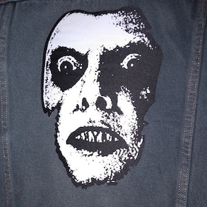 May include: A black and white image of a face with a wide open mouth and large eyes. The face is printed on a dark blue denim jacket.