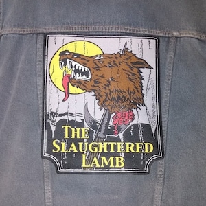 May include: A grey denim vest with a large patch depicting a snarling wolf with a red tongue against a yellow moon. The patch reads "THE SLAUGHTERED LAMB" in yellow text. The vest has button closures and visible stitching.