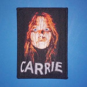 May include: A black and white embroidered patch with a portrait of Carrie White from the movie Carrie. The patch has the word "CARRIE" embroidered in white letters below the portrait.