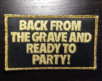 Puede incluir: Parche rectangular negro con texto dorado que dice "BACK FROM THE GRAVE AND READY TO PARTY!". El parche tiene un borde dorado y está sobre una superficie de madera oscura.