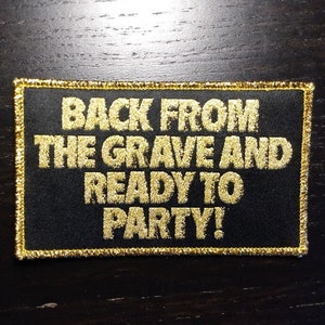 Puede incluir: Parche rectangular negro con texto dorado que dice "BACK FROM THE GRAVE AND READY TO PARTY!". El parche tiene un borde dorado y está sobre una superficie de madera oscura.
