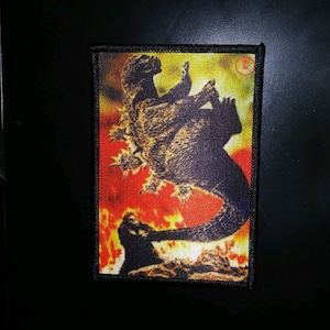 May include: A black and white embroidered patch featuring a Godzilla-like monster with a red and yellow background. The monster is standing on a rock with flames behind it.