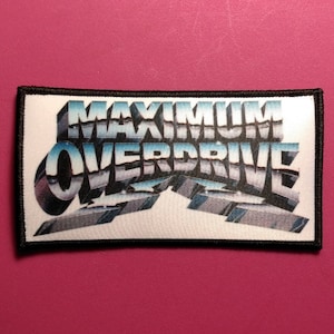 以下が含まれることがあります： 「MAXIMUM OVERDRIVE」の文字が3Dクロム風フォントで描かれた長方形のパッチ。青と白のグラデーションの背景に配置されています。パッチは黒い縁取りがあり、ピンク色の表面に置かれています。