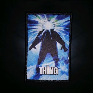 May include: A blue and white embroidered patch featuring a silhouette of a creature from the film "The Thing" and the word "THING" in white text.