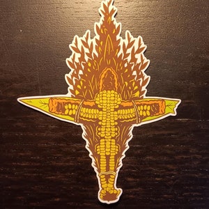 May include: A yellow corn cob with brown husks is arranged in the shape of a person with outstretched arms. The corn cob is surrounded by brown and green leaves.