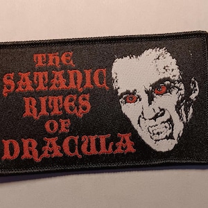 Puede incluir: Parche rectangular negro con el texto "THE SATANIC RITES OF DRACULA" en rojo. Ilustración en blanco y negro de Drácula con ojos rojos.