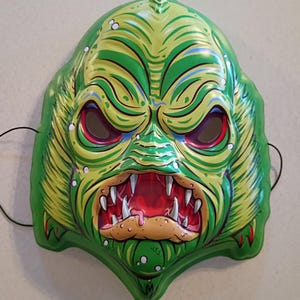 May include: A green and yellow monster mask with red eyes and sharp white teeth. The mask has a menacing expression and is made of a shiny material. Black string is attached for wearing.