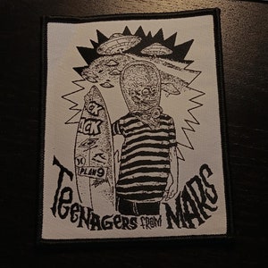 May include: Black and white embroidered patch featuring a cartoon alien holding a surfboard with the text "Plan 9" and "Teenage from Mars".