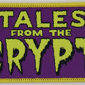 May include: A rectangular embroidered patch with a purple background and a yellow stitched border. The text TALES FROM THE CRYPT is displayed in a lime green, dripping font with black drop shadows, centered on the purple fabric surface.