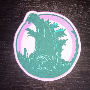 May include: A green and pink embroidered patch featuring a silhouette of Godzilla standing in front of a city skyline.