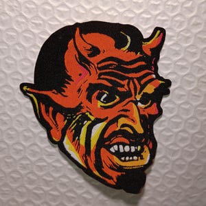 May include: A patch featuring a stylized devil's head with orange, yellow, and black colors. The devil has horns, a goatee, and a menacing expression. The patch is likely for clothing or accessories.