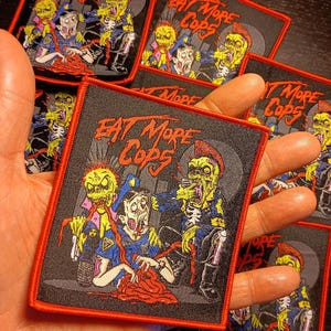 May include: A black and red embroidered patch featuring a graphic of three zombie-like figures with red blood splatters. The text "EAT MORE COPS" is in red and white lettering.