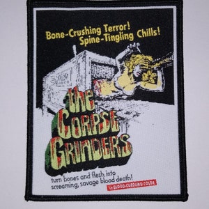 May include: A white and black embroidered patch with a graphic of a woman being pulled into a machine. The text reads "Bone-Crushing Terror! Spine-Tingling Chills! The Corpse Grinders turn bones and flesh into screaming, savage blood death! In Blood-Curdling Color."