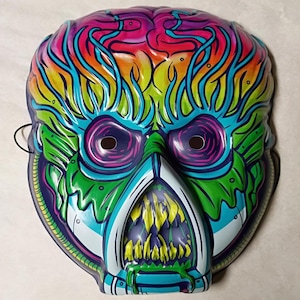Op de afbeelding: Een levendig, kleurrijk masker met een grotesk ontwerp. Het masker heeft een hersenachtig bovenstuk in roze, blauw en oranje, met groene en blauwe gelaatstrekken. De mond heeft gele tanden. Het masker heeft ooggaten en een zwart touwtje.