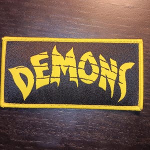 May include: Yellow embroidered patch with the word "DEMONS" in a stylized font on a black background.