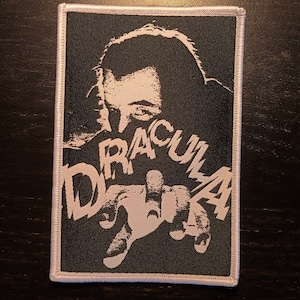 May include: Black and white embroidered patch with a stylized image of a person's face and hand with the word "DRACULA" written in a bold, stylized font.