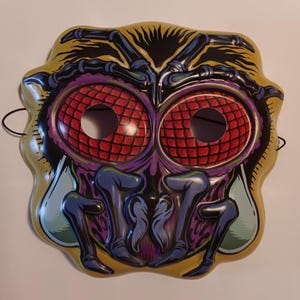 May include: A Halloween mask with a bug-like design. The mask features large, red, grid-patterned eyes, purple and blue accents, and a yellow and black outer edge. The mask has eye holes and a black string for wearing.