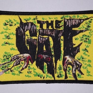 May include: A yellow and black embroidered patch with the text "THE GATE" in black. The text is surrounded by green foliage and two black hands reaching out from the sides.