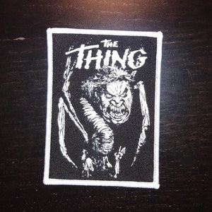 Może przedstawiać: Czarno-biała naszywka haftowana z białym obramowaniem. Naszywka przedstawia stworzenie z ostrymi zębami i pazurami oraz tekstem "THE THING" nad nim.