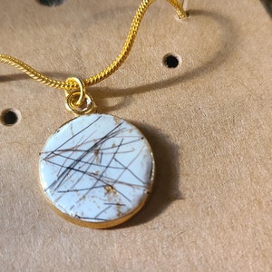 Peut inclure: Un collier en chaîne dorée avec un pendentif rond orné d'une pierre blanche avec des inclusions noires et dorées.