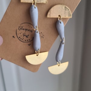 Puede incluir: Un par de pendientes colgantes con formas de lágrima de arcilla gris y acentos de medio círculo dorado. Los pendientes se muestran en una tarjeta marrón con el texto "Blackgrass Girl Designs".